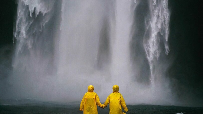 Zwei Menschen stehen mit knallgelben Regenmänteln händehaltend vor einem tosenden Wasserfall. 