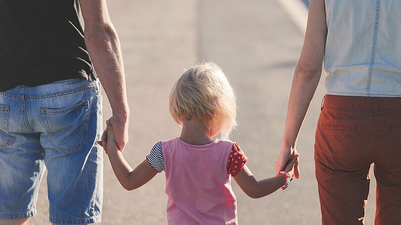 Ein Vater, ein Kind und eine Mutter gehen Hand in Hand. Man sieht sie von hinten. Der Vater träge eine knielange Jeans und ein blaues T-Shirt, das Kind ein rosa T-Shirt und die Mutter eine rote Jeans und ein weißes T-Shirt. 