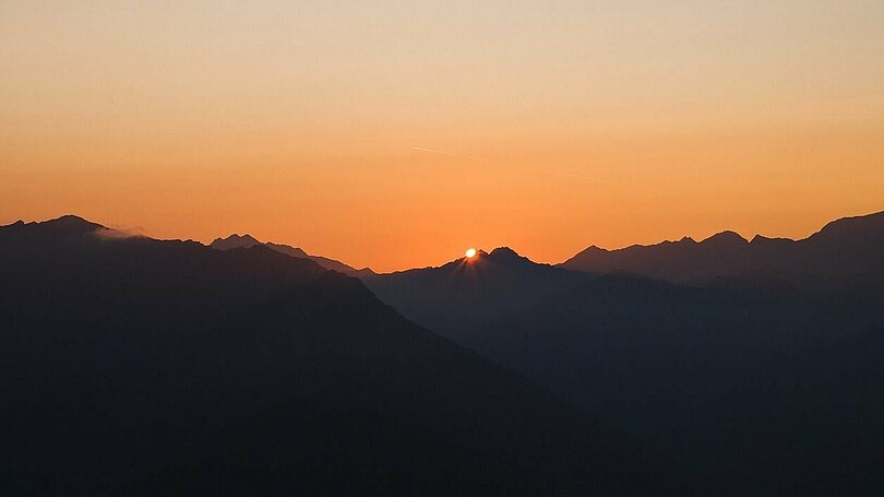 Abenstimmung in den Tiroler Bergen: Dunkle Bergketten, oranger Himmel und die Sonne als kleiner, gelber Punkt in der Mitte des Bildes und am Rand der Berge.