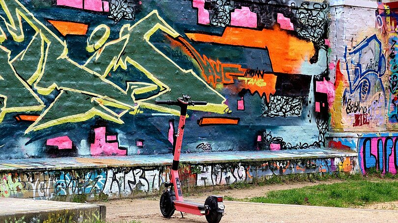 Street Art: Graffity auf einer Mauer mit orange, pink, blau, grün und gelb. Ein rosa E-Scooter steht davor. 