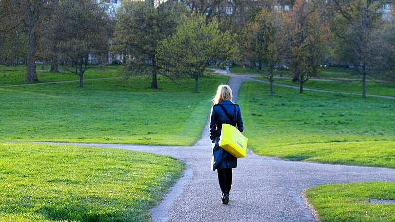 Eien Frau in dunklem Mantel und mit gelber Tasche geht in einem Park auf eine Kreuzug zu. 