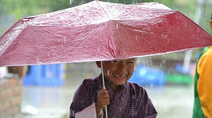 Ein asiatisch aussehender Junge in lila Gewand mit weißen Ärmeln steht lachend unter einem rosa Regenschirm, während es regnet. 