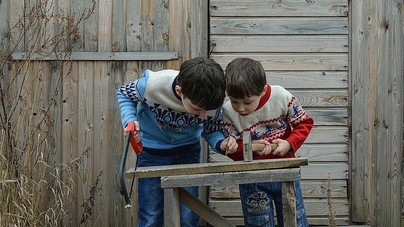 Zwei Buben mit bunten Pullovern stehen vor einer grauen Holzwand und suchen einen geeigneten Platz um einen Nagel in zwei Bretter, die auf Schragen liegen,  einzuschlagen