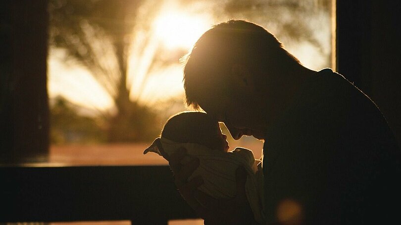 Ein Vater schaut sein neugeborenes Kind an und berührt mit seiner Stirn, die Stirn des Kindes. Im Hintergrund geht die Sonne hinter Palmen unter. Von den Figuren im Vordergrund sieht man die dunklen Umrisse. 